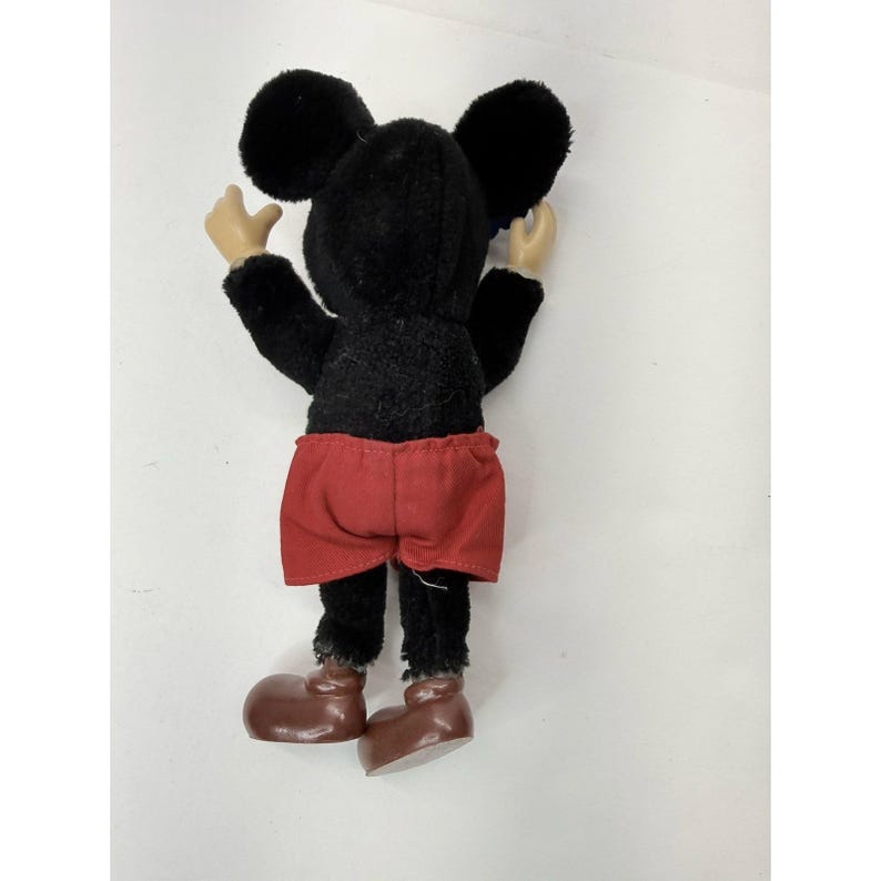 May include: A vintage Mickey Mouse plush toy, viewed from the back. The plush features a black body, large round ears, red shorts, and brown shoes. The arms and hands are a light tan color. The toy is standing with its arms raised.