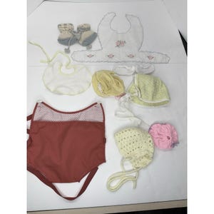 May include: A collection of baby items, including a red baby carrier, bibs, booties, and bonnets. The bibs are white with floral embroidery. The booties are blue and beige. The bonnets are yellow and green. The baby carrier is red with a white polka dot pattern.