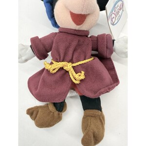 May include: A plush Mickey Mouse doll wearing a burgundy outfit with a yellow rope belt. The doll has white gloves, brown shoes, and a black and blue hat. A Disney Store tag is attached.