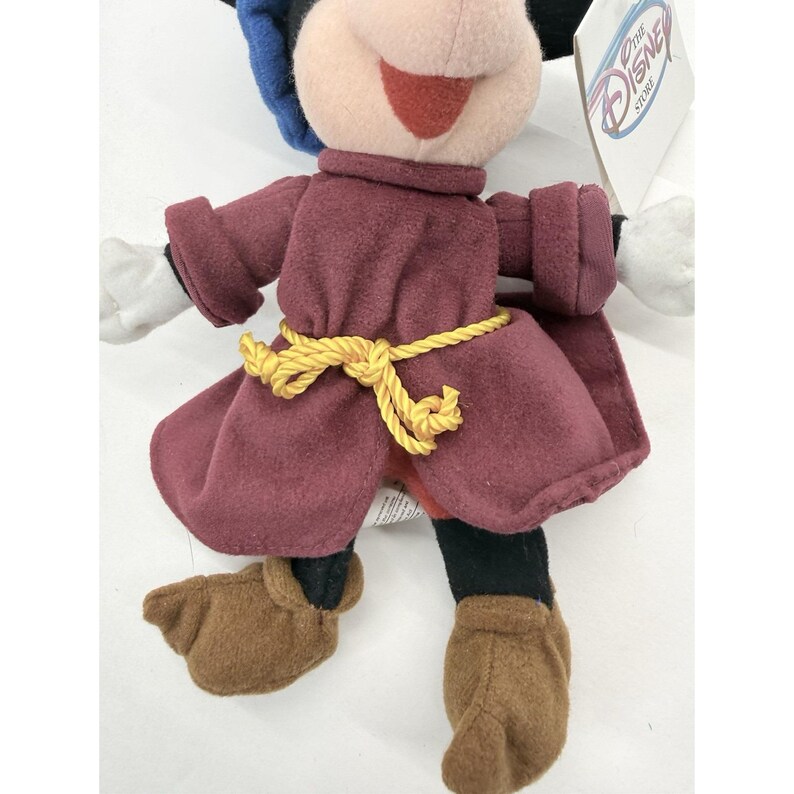 May include: A plush Mickey Mouse doll wearing a burgundy outfit with a yellow rope belt. The doll has white gloves, brown shoes, and a black and blue hat. A Disney Store tag is attached.