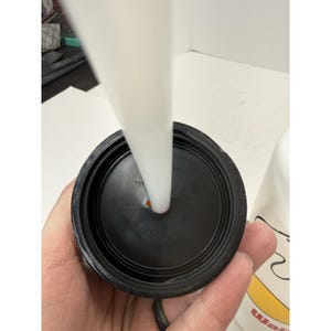 May include: Close-up of a black plastic lid with a white straw inserted. The lid has a circular design with a small hole for the straw. The straw is white and cylindrical. The lid is held by a hand.