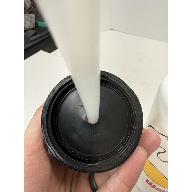 May include: Close-up of a black plastic lid with a white straw inserted. The lid has a circular design with a small hole for the straw. The straw is white and cylindrical. The lid is held by a hand.