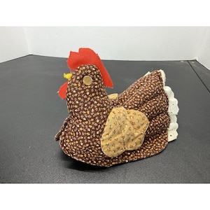 May include: A fabric chicken figurine featuring a brown floral print body, a bright red felt comb, a yellow beak, and a single button eye. The wing is made of a lighter tan floral fabric, and the tail consists of white eyelet lace trim.