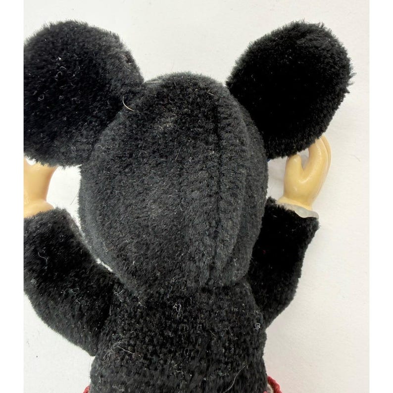 May include: A vintage Mickey Mouse plush toy, viewed from the back, with large black ears and a black fuzzy body. The toy has tan arms and hands, and a red and white patterned detail is visible at the bottom.