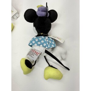 K&ouml;nnte beinhalten: Minnie Mouse Pl&uuml;schtier mit schwarzen Ohren, einem lila Hut und einem blauen Kleid mit wei&szlig;en Punkten. Das Spielzeug hat gelbe Schuhe und wei&szlig;e Handschuhe. Ein Etikett des Herstellers GUND ist angebracht.
