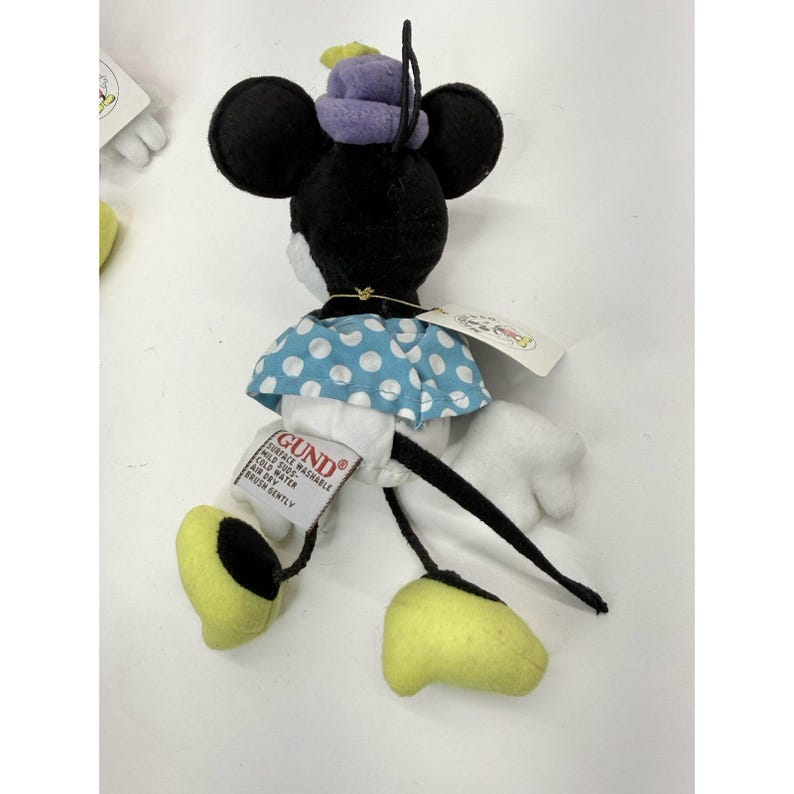 K&ouml;nnte beinhalten: Minnie Mouse Pl&uuml;schtier mit schwarzen Ohren, einem lila Hut und einem blauen Kleid mit wei&szlig;en Punkten. Das Spielzeug hat gelbe Schuhe und wei&szlig;e Handschuhe. Ein Etikett des Herstellers GUND ist angebracht.