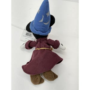 May include: A Sorcerer Mickey Mouse bean bag plush toy. The plush is wearing a blue wizard hat with silver stars and moons, a burgundy robe, and brown shoes. The toy has black ears and white gloves. A tag is attached.