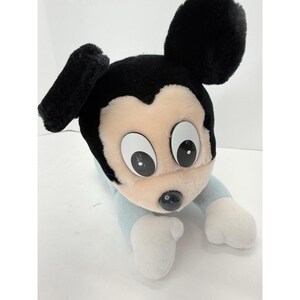 Puede incluir: Un peluche de un personaje de dibujos animados con ojos grandes y redondos y una nariz negra. El juguete tiene orejas y pelo negros, un cuerpo azul claro y manos blancas. El juguete est&aacute; tumbado.