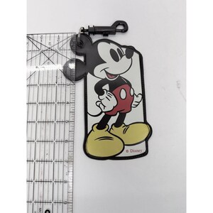 May include: A Mickey Mouse luggage tag with a black clip. The tag features a smiling Mickey Mouse in his classic outfit: black ears, white face, red shorts, yellow shoes, and white gloves. The tag is approximately 4 inches tall.