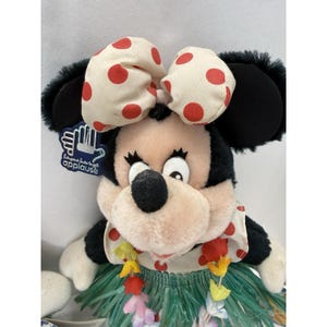 Puede incluir: Una mu&ntilde;eca de peluche de Minnie Mouse con un atuendo hawaiano. La mu&ntilde;eca tiene un lazo color crema con lunares rojos, orejas negras y una falda de hierba. Una etiqueta dice "Everyone loves to applaud".