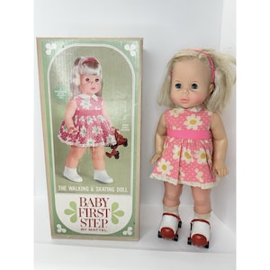 May include: A vintage Baby First Step doll by Mattel, with blonde hair and blue eyes, wearing a pink floral dress and roller skates. The doll stands next to its original box, which features an illustration of the doll. The box reads "The Walking & Skating Doll".