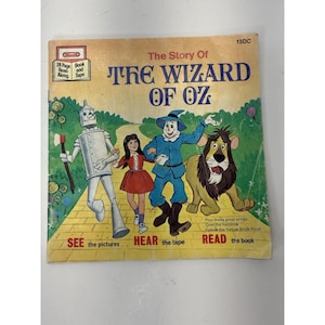 May include: Vintage "The Wizard of Oz" book and tape set. The cover features illustrations of the Tin Man, Dorothy, the Scarecrow, and the Cowardly Lion walking on a yellow brick road. The text reads "The Story Of THE WIZARD OF OZ."