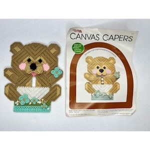 May include: A brown and beige plastic canvas craft kit featuring a baby bear design. The kit includes pre-cut plastic canvas shapes and instructions for creating a switchplate cover. The packaging includes the text "Canvas Capers" and "Designed for your pleasure by Dick Martin".