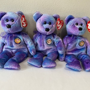 May include: Three purple and blue tie-dye beanie babies with a gold star and Ty logo on their chests. The beanie babies are sitting on a white surface.