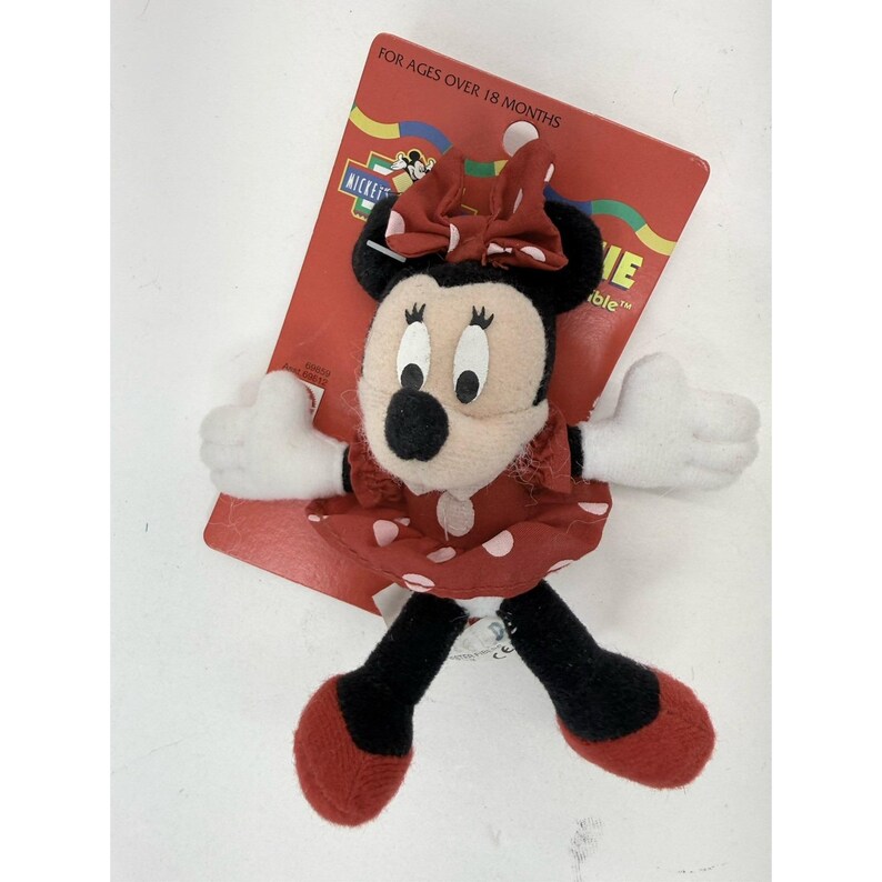 Puede incluir: Una mu&ntilde;eca de peluche de Minnie Mouse con un vestido y un lazo rojos, ambos con lunares blancos. La mu&ntilde;eca tiene orejas negras, una nariz negra y guantes blancos. La mu&ntilde;eca est&aacute; sujeta a una etiqueta de cart&oacute;n roja que dice "FOR AGES OVER 18 MONTHS."