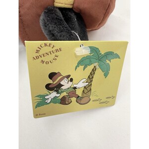 K&ouml;nnte beinhalten: Ein gelbes Etikett mit der Aufschrift "Mickey Adventure Mouse" und einer Cartoon-Illustration von Mickey Mouse in Safari-Kleidung. Mickey tr&auml;gt einen braunen Hut und khakifarbene Kleidung. Eine gr&uuml;ne Palme ist ebenfalls abgebildet.
