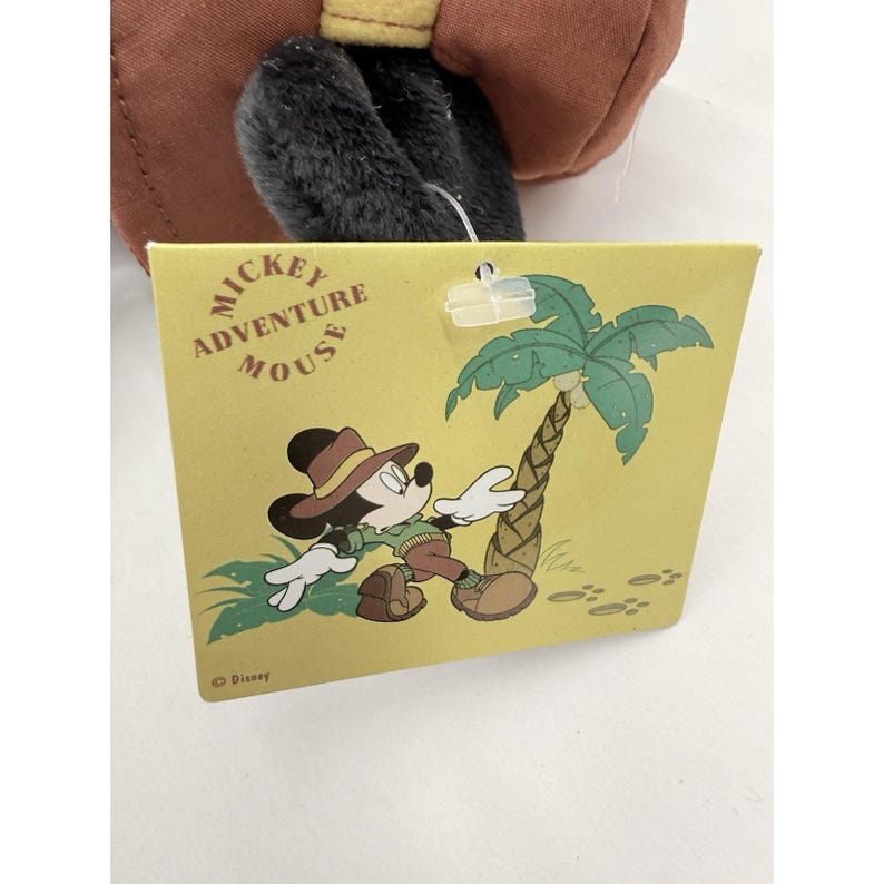 K&ouml;nnte beinhalten: Ein gelbes Etikett mit der Aufschrift "Mickey Adventure Mouse" und einer Cartoon-Illustration von Mickey Mouse in Safari-Kleidung. Mickey tr&auml;gt einen braunen Hut und khakifarbene Kleidung. Eine gr&uuml;ne Palme ist ebenfalls abgebildet.