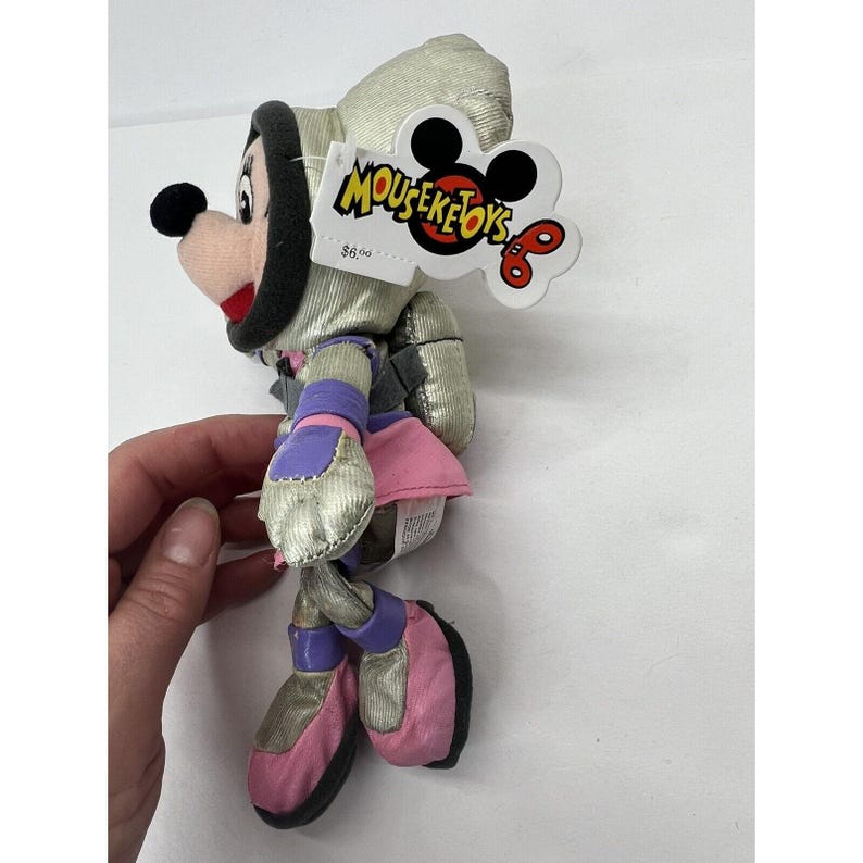 May include: A silver and pink plush toy of a mouse wearing a space suit. The toy has a purple and silver belt and pink boots. The toy has a tag that says "MousekeToys" with a price tag of $6.00.