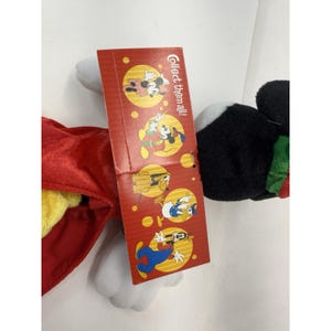 Puede incluir: Un peluche de Mickey Mouse con una capa roja y una etiqueta que dice "&iexcl;Colecci&oacute;nalos todos!". La etiqueta presenta ilustraciones de Mickey, Minnie, Goofy y el Pato Donald. El juguete tiene detalles en negro, blanco y amarillo.