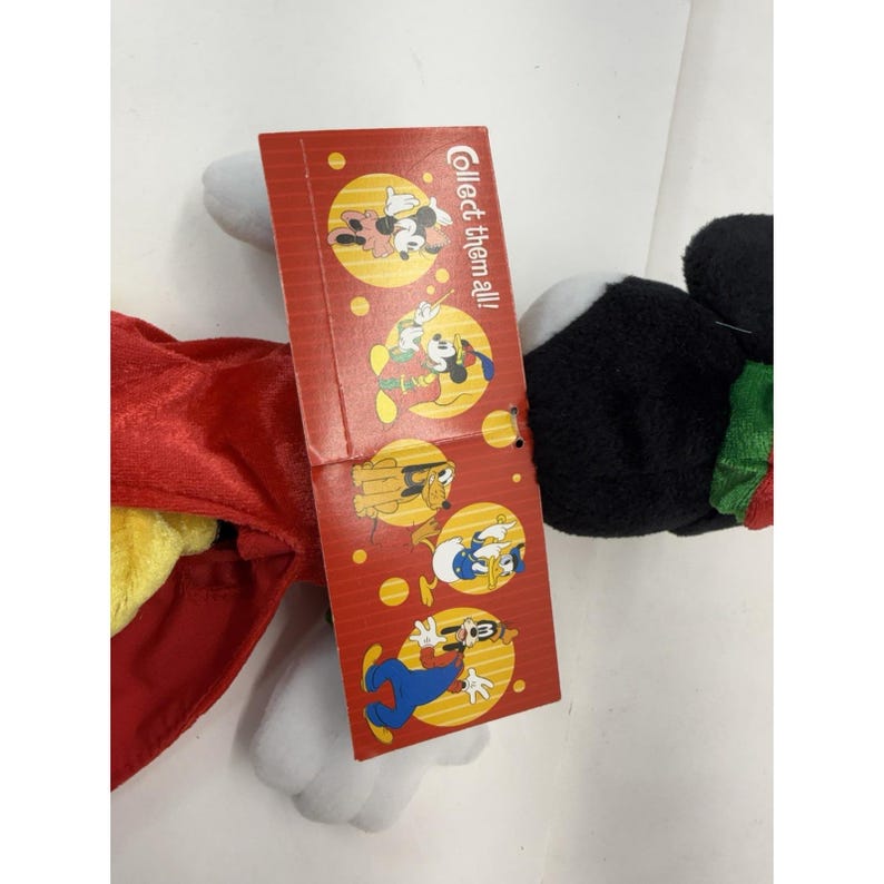 Puede incluir: Un peluche de Mickey Mouse con una capa roja y una etiqueta que dice "&iexcl;Colecci&oacute;nalos todos!". La etiqueta presenta ilustraciones de Mickey, Minnie, Goofy y el Pato Donald. El juguete tiene detalles en negro, blanco y amarillo.