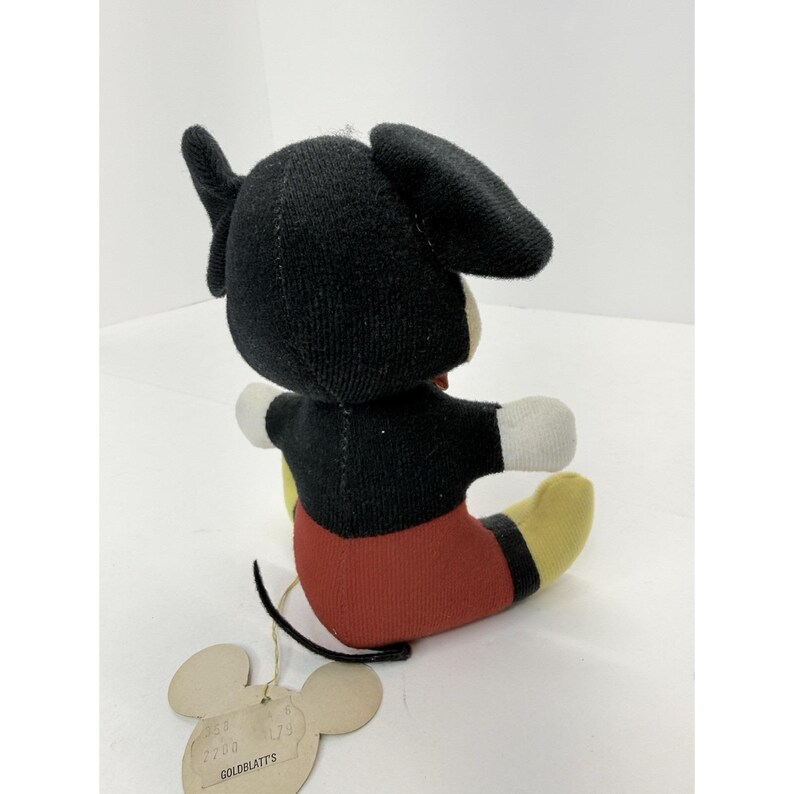 Puede incluir: Un peluche vintage de Mickey Mouse, con cabeza y orejas negras, torso negro, pantalones cortos rojos, pies amarillos y guantes blancos. El juguete tiene una etiqueta que dice "Goldblatt's". El peluche est&aacute; sentado.