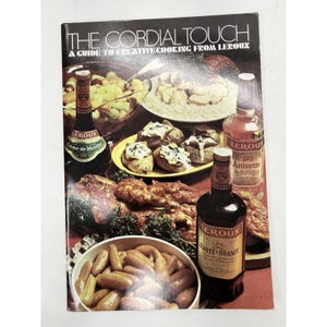 May include: A vintage cookbook cover titled "The Cordial Touch" with the subtitle "A Guide to Creative Cooking From Leroux." The cover features various dishes and bottles of Leroux liqueurs, including Creme de Menthe, Anisette, and Coffee Brandy.