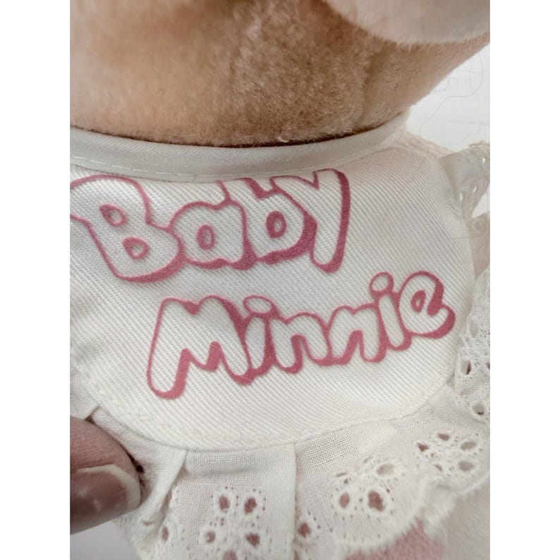 Puede incluir: Primer plano del cuello de una mu&ntilde;eca de peluche. El cuello blanco presenta el texto "Baby Minnie" en escritura rosa. El cuello est&aacute; adornado con un delicado encaje de ojal.