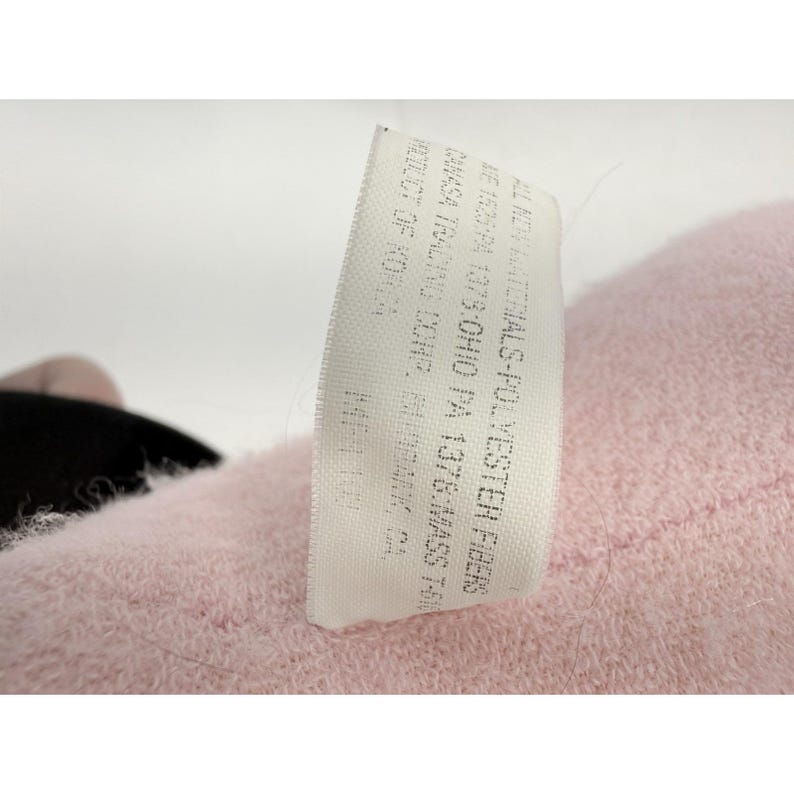 Puede incluir: Primer plano de un juguete de peluche rosa con una etiqueta blanca. La etiqueta contiene texto como "ALL NEW MATERIALS-POLYESTER FIBERS". El juguete tiene una textura suave y difusa.