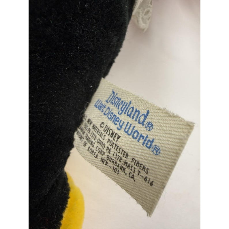 May include: A close-up of a tag from a Disneyland Walt Disney World plush toy. The tag is rectangular with rounded corners and features the Disneyland and Walt Disney World logos in blue. The tag also includes text about the materials and manufacturer.