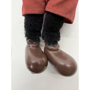 May include: Close-up of a doll's legs. The doll's legs are covered in black, fuzzy material, and the feet are brown, rounded, and shiny. The doll is wearing red pants.