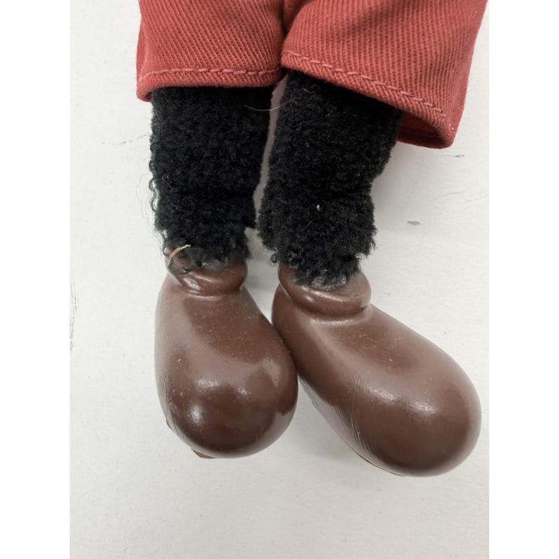 May include: Close-up of a doll's legs. The doll's legs are covered in black, fuzzy material, and the feet are brown, rounded, and shiny. The doll is wearing red pants.