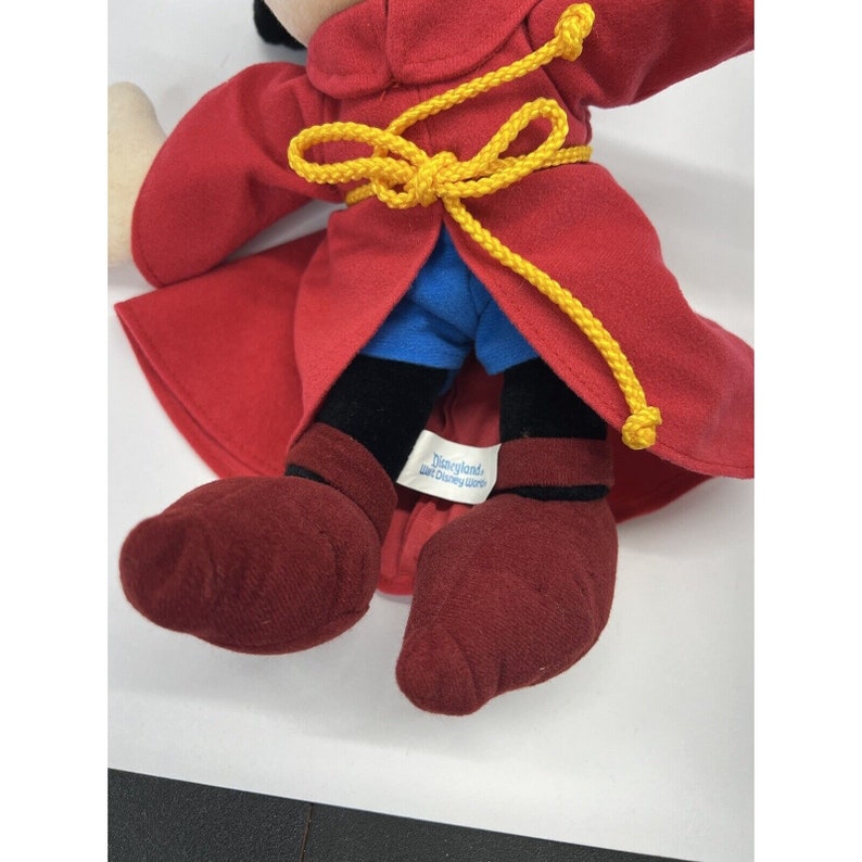 May include: Red plush Mickey Mouse doll wearing a red robe with a yellow rope tied around the waist. The doll has red shoes and a blue shirt. The doll has a Disneyland label on the back of the robe.