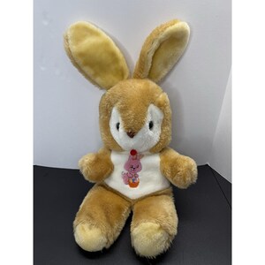 May include: A plush stuffed rabbit with tan fur, long upright ears, and a white face patch. The rabbit features a white chest panel with an embroidered pink bunny graphic holding a colorful basket, accented by a small red pom-pom at the neck.