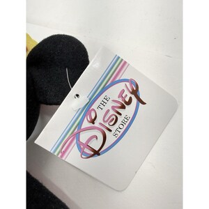 Puede incluir: Un juguete de peluche negro con un acento amarillo es parcialmente visible junto a una etiqueta blanca. La etiqueta presenta el logotipo de Disney Store en un &oacute;valo rosa y azul con las palabras "THE DISNEY STORE" en una fuente de escritura.