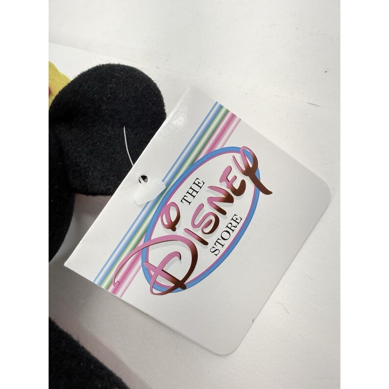 Puede incluir: Un juguete de peluche negro con un acento amarillo es parcialmente visible junto a una etiqueta blanca. La etiqueta presenta el logotipo de Disney Store en un &oacute;valo rosa y azul con las palabras "THE DISNEY STORE" en una fuente de escritura.