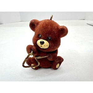 May include: A brown plush teddy bear ornament playing a triangle. The bear has a brown nose and black eyes.