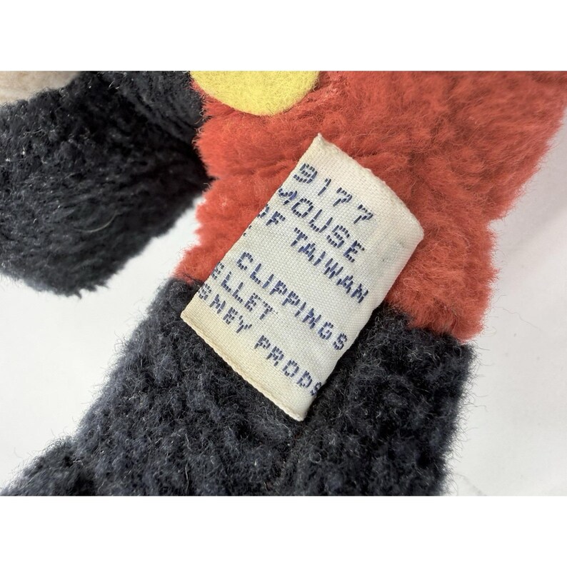 May include: Close-up of a vintage plush toy with black, red, and yellow accents. A white tag is attached, displaying text including "9177 HOUSE OF TAIWAN CLIPPINGS DISNEY PRODS."