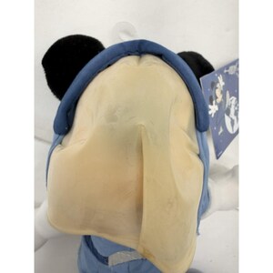 May include: A plush toy of a cartoon character, featuring a large, light yellow face, black ears, and a blue hat. The toy is wearing a blue outfit. A tag with a cartoon graphic is visible.
