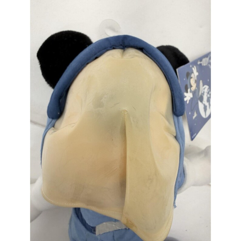 May include: A plush toy of a cartoon character, featuring a large, light yellow face, black ears, and a blue hat. The toy is wearing a blue outfit. A tag with a cartoon graphic is visible.