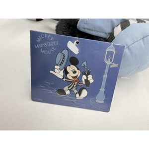 May include: A Mickey Mouse plush toy with a tag featuring a cartoon illustration of Mickey Mouse in a pinstripe suit, holding a hat and umbrella. The tag reads "Mickey Mainstreet Mouse" in white text on a blue background.
