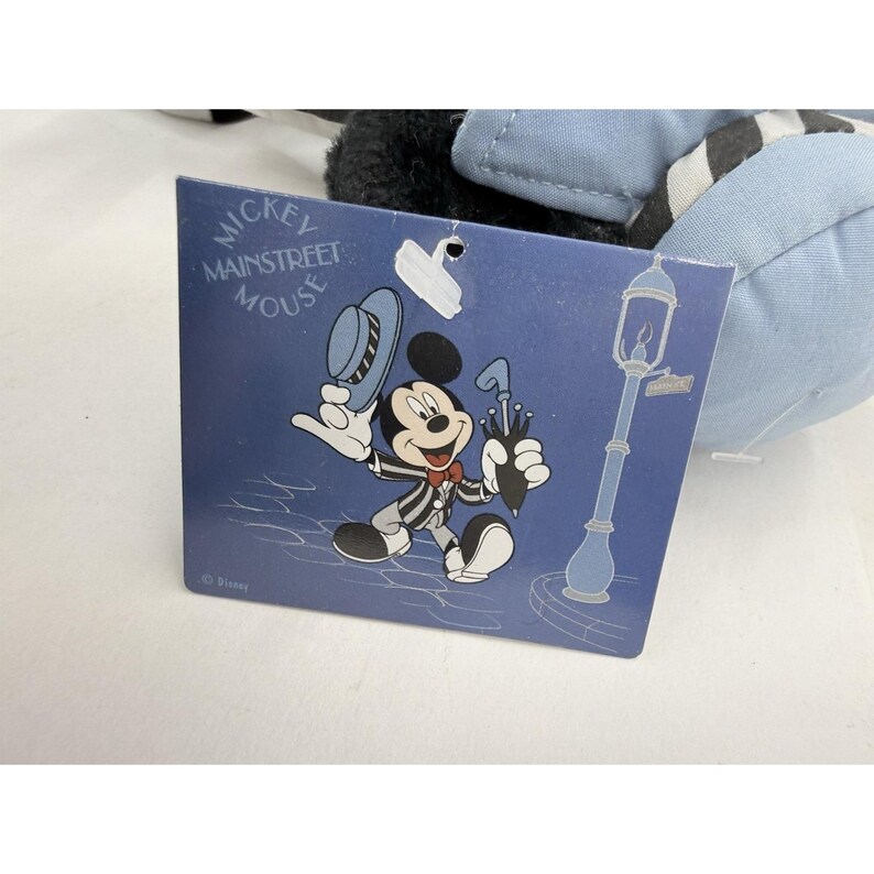 May include: A Mickey Mouse plush toy with a tag featuring a cartoon illustration of Mickey Mouse in a pinstripe suit, holding a hat and umbrella. The tag reads "Mickey Mainstreet Mouse" in white text on a blue background.