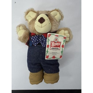 May include: A brown teddy bear wearing a red and blue plaid shirt, a blue bow tie with white polka dots, blue denim overalls, and brown boots. The bear is holding a tag that says "Wendy's Old Fashioned Hamburgers" with a picture of Wendy.