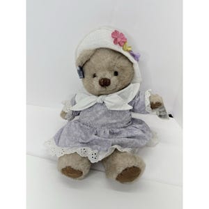 May include: A plush teddy bear wearing a lavender floral print dress with white lace trim and a matching bonnet adorned with flowers. The bear has a brown nose and is seated.