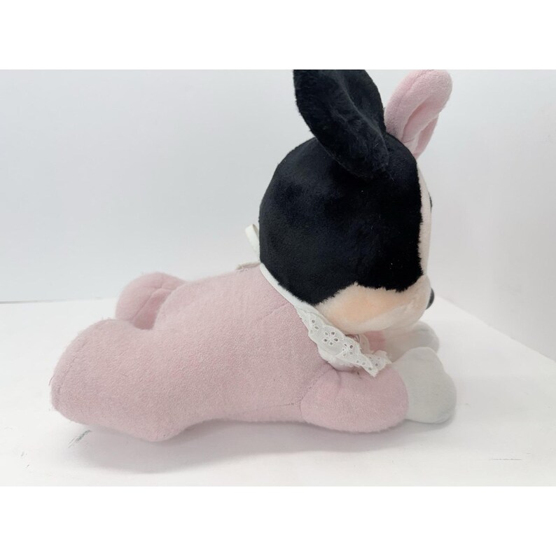 Puede incluir: Un peluche con forma de conejito, con cabeza y orejas negras, interior de las orejas rosa y cuerpo rosa. El juguete tiene un cuello de encaje blanco y est&aacute; tumbado.