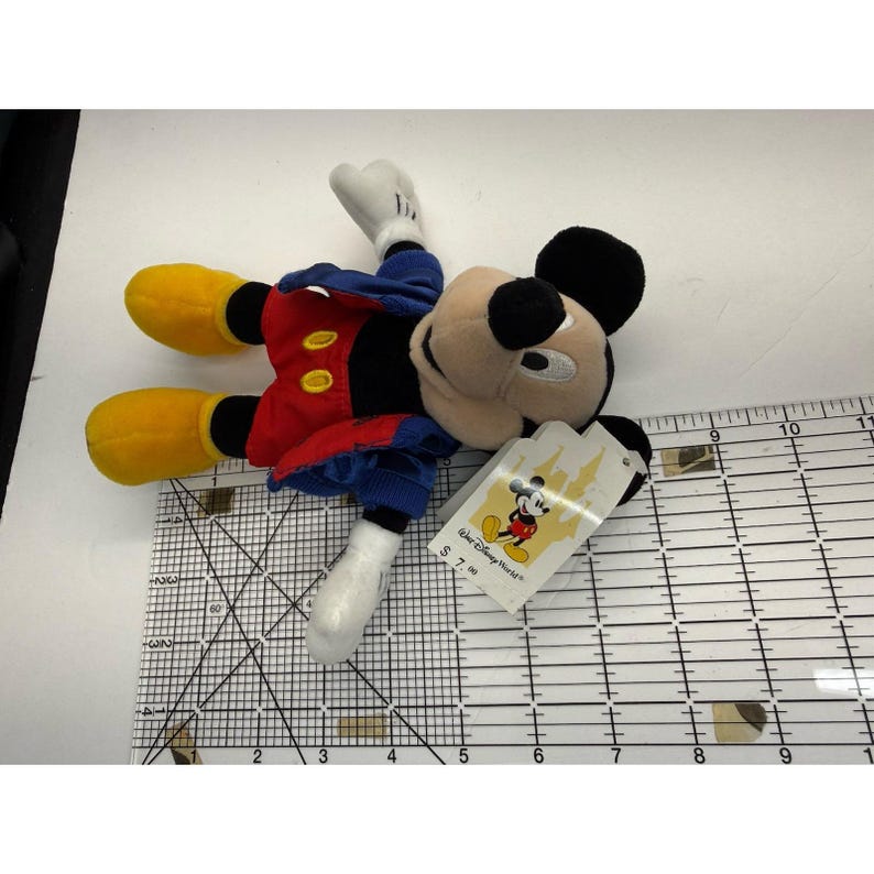 Peut inclure: Une peluche de Mickey Mouse, allong&eacute;e sur le dos. Mickey porte un short rouge avec des boutons jaunes, une veste bleue, des chaussures jaunes et des gants blancs. Une &eacute;tiquette Disney World est attach&eacute;e.