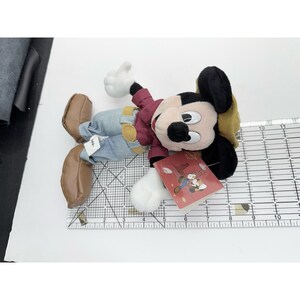 K&ouml;nnte beinhalten: Ein Mickey Mouse Pl&uuml;schtier, das auf der Seite liegt. Das Spielzeug tr&auml;gt eine bordeauxfarbene Jacke, blaue Jeans und braune Schuhe. Es hat schwarze Ohren, ein beiges Gesicht und wei&szlig;e Handschuhe. Ein rotes Etikett mit einer Illustration ist angebracht.