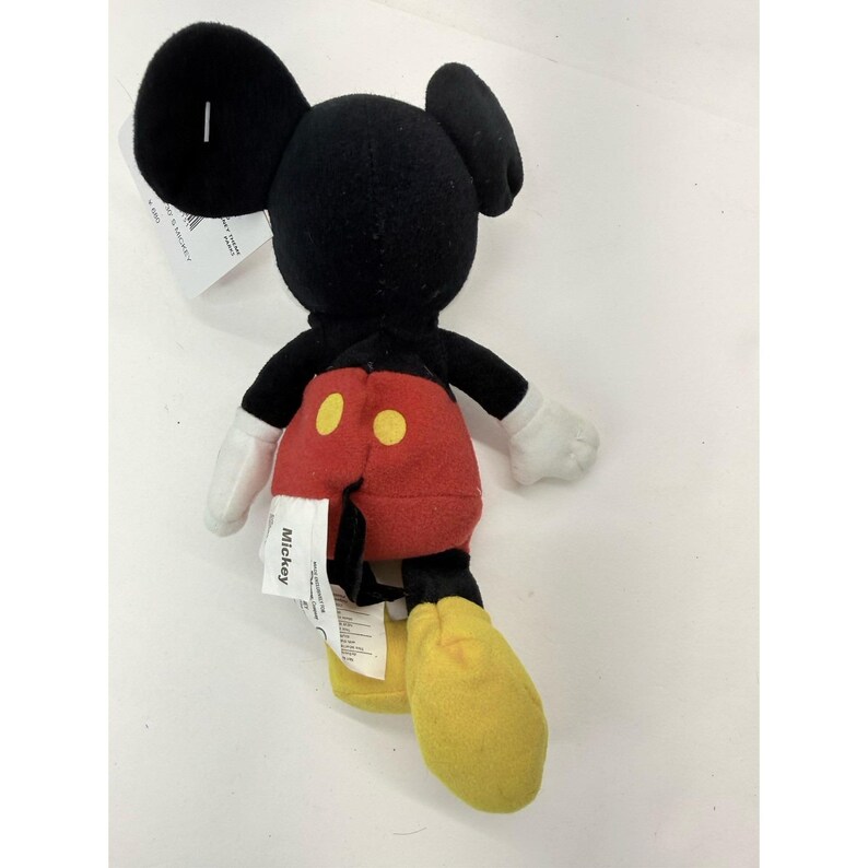 Puede incluir: Un peluche de Mickey Mouse, visto desde atr&aacute;s. El juguete presenta una cabeza y orejas negras, pantalones cortos rojos con botones amarillos, guantes blancos y zapatos amarillos. Una etiqueta blanca con el nombre "Mickey" est&aacute; adjunta.