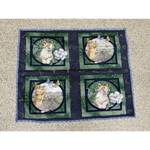 May include: A rectangular fabric quilt featuring four square panels with dark blue borders. Each panel displays a circular illustration of cats in green foliage with white flowers and butterflies. The quilt has a blue patterned edge trim.