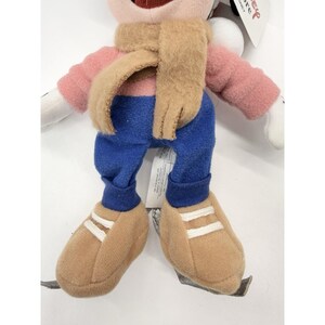 May include: A plush toy character wearing a pink shirt, blue pants, and tan shoes with white stripes. It has a tan scarf around its neck. The toy has white gloves and a smiling face.