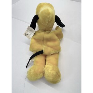 Puede incluir: Un juguete de peluche con forma de perro amarillo con orejas y cola negras. El juguete tiene una etiqueta adjunta. El perro est&aacute; en posici&oacute;n sentada, con las patas extendidas. El juguete es de material suave.